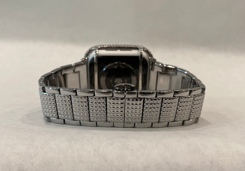 May include: A silver-toned smartwatch with a matching band. The watch face is encrusted with small, sparkling stones. The band features a textured, patterned design, and the watch is set against a plain background.