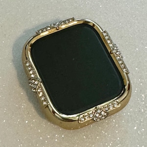 May include: A gold-colored watch case with a black face and a diamond-like trim. The case is designed to fit a smart watch.