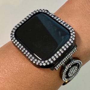 May include: A black smartwatch case featuring a square frame encrusted with clear, sparkling rhinestones. The watch band is also adorned with matching rhinestones in a circular link pattern, creating a cohesive, decorative appearance on the wrist.