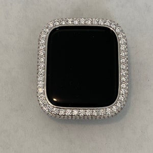 May include: A silver-colored watch case with a black screen, encrusted with small, clear gemstones around the bezel. The watch case has a rectangular shape with rounded corners, and the gemstones are closely set.