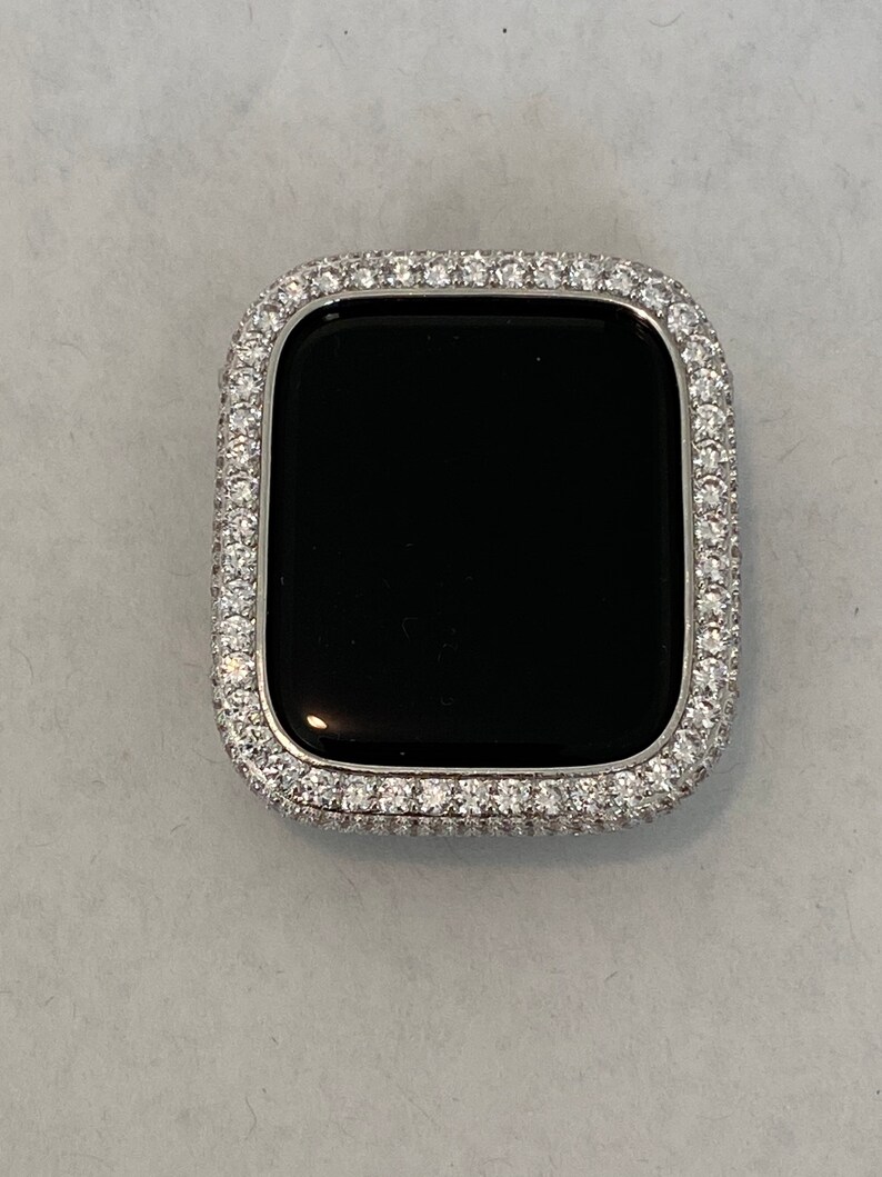 May include: A silver-colored watch case with a black screen, encrusted with small, clear gemstones around the bezel. The watch case has a rectangular shape with rounded corners, and the gemstones are closely set.
