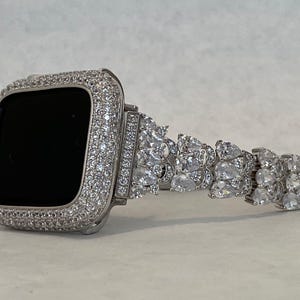 May include: A silver Apple Watch band with a diamond-like design. The band is made of metal and has a clear, sparkly finish.