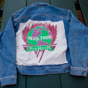 Pode incluir: Uma jaqueta de ganga azul com um remendo branco nas costas. O remendo apresenta um globo terrestre com chamas ao redor e o texto "1979 TOUR VAN HALEN".