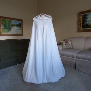 May include: A white hooded cape with a long flowing design. The cape has a single button closure at the neck and a small decorative detail on the hood.