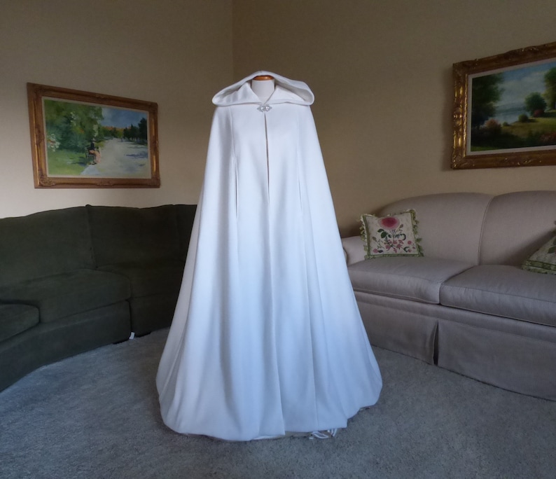 May include: A white hooded cape with a long flowing design. The cape has a single button closure at the neck and a small decorative detail on the hood.