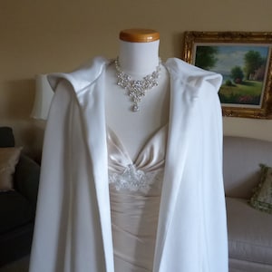 May include: A white satin bridal cape with a hood, worn over a white dress with a lace bodice and a sparkling rhinestone necklace.