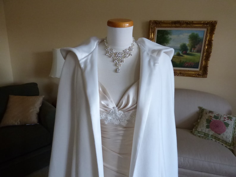 May include: A white satin bridal cape with a hood, worn over a white dress with a lace bodice and a sparkling rhinestone necklace.