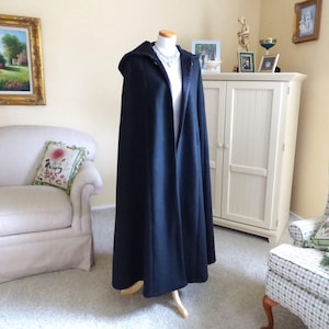 May include: A long black hooded cape with a zipper closure. The cape is displayed on a mannequin in a room with furniture.