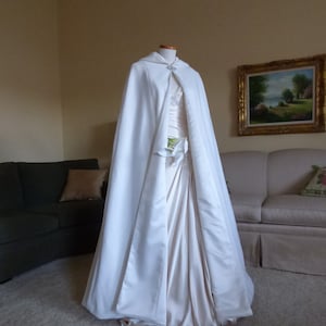 May include: A white satin bridal cape with a hood and a small jeweled brooch. The cape is draped over a white satin dress.