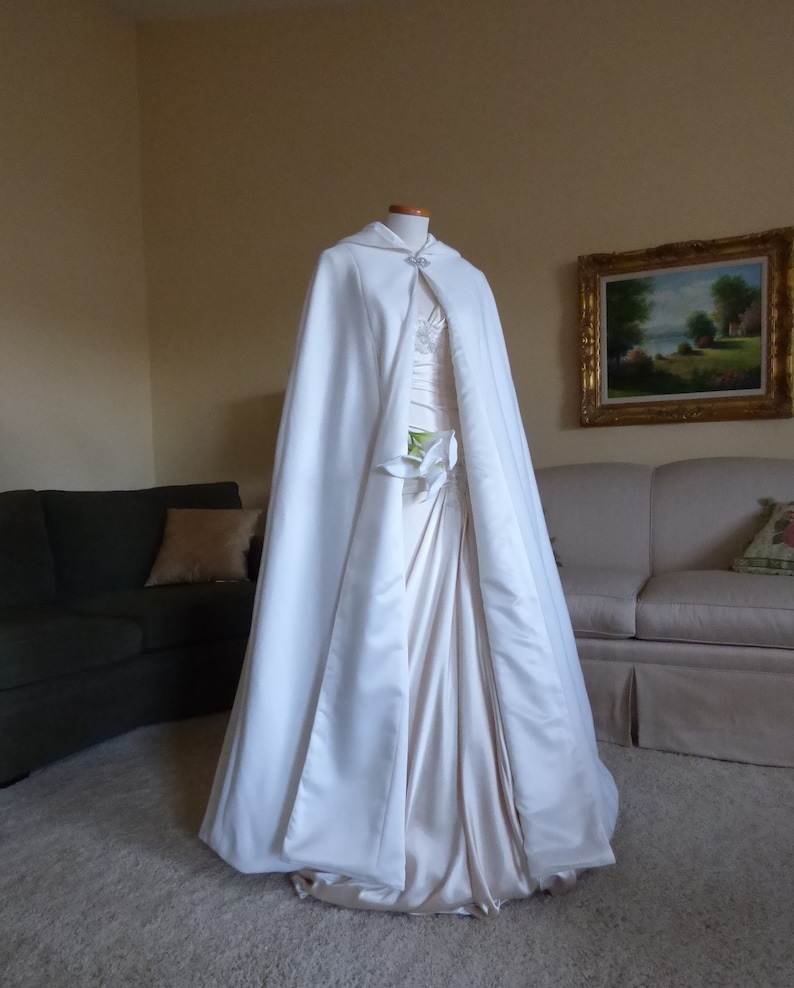 May include: A white satin bridal cape with a hood and a small jeweled brooch. The cape is draped over a white satin dress.