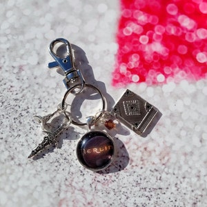 Puede incluir: Un llavero plateado con un peque&ntilde;o amuleto de libro, un amuleto de espada, un amuleto de vidrio con la palabra "BERLIN" en &eacute;l y un anillo de llave plateado.