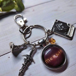 Puede incluir: Un llavero de plata con un dije que dice "Merlin" en letras doradas sobre un fondo morado oscuro. El llavero tambi&eacute;n incluye un dije de espada de plata, un dije de libro de plata y un dije de trompeta de plata.