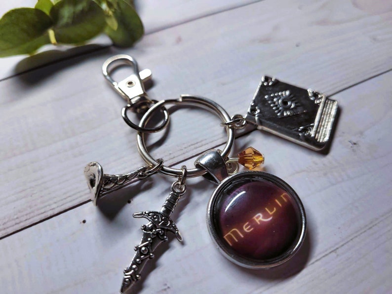 Puede incluir: Un llavero de plata con un dije que dice "Merlin" en letras doradas sobre un fondo morado oscuro. El llavero tambi&eacute;n incluye un dije de espada de plata, un dije de libro de plata y un dije de trompeta de plata.