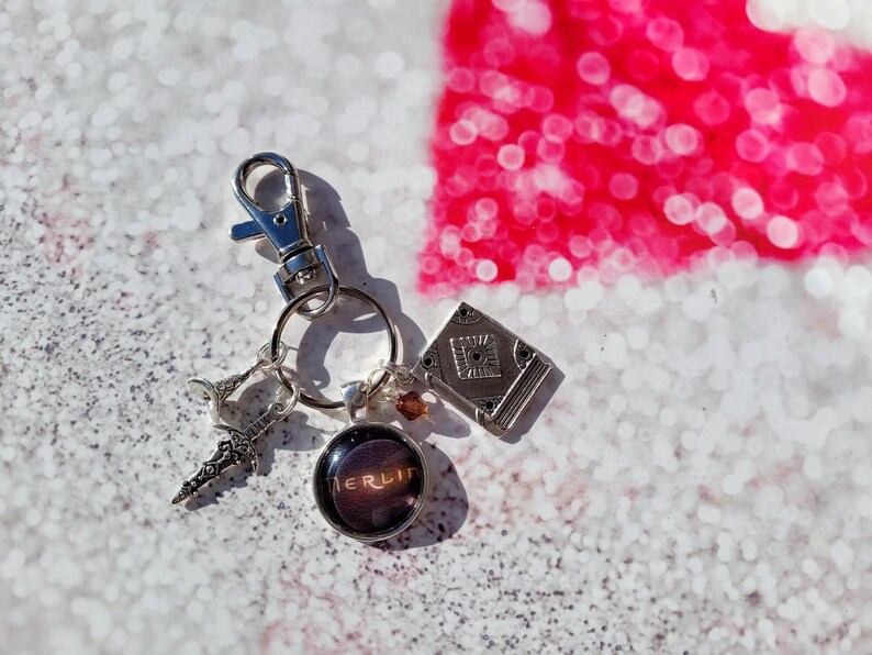 Puede incluir: Un llavero plateado con un peque&ntilde;o amuleto de libro, un amuleto de espada, un amuleto de vidrio con la palabra "BERLIN" en &eacute;l y un anillo de llave plateado.