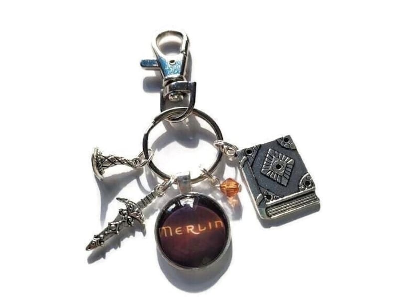 Puede incluir: Un llavero de plata con un dije que dice "Merlin" en rojo oscuro y naranja. El llavero tambi&eacute;n tiene una peque&ntilde;a espada de plata, un dije de libro de plata y un peque&ntilde;o dije de corona de plata.