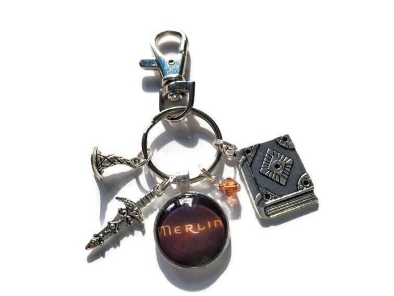 Puede incluir: Un llavero de plata con un dije que dice "Merlin" en color rojo oscuro y naranja. El llavero tambi&eacute;n tiene un dije de un libro, una espada y un peque&ntilde;o objeto de plata.