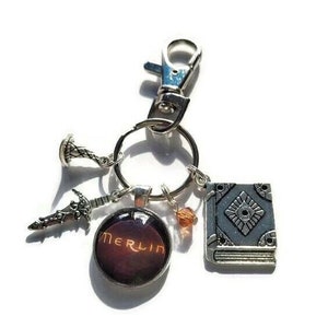 Puede incluir: Un llavero plateado con un dije que dice "Merlin" en una fuente roja y naranja. El llavero tambi&eacute;n tiene un dije de libro plateado, un dije de espada y un peque&ntilde;o dije plateado con un dise&ntilde;o en espiral.