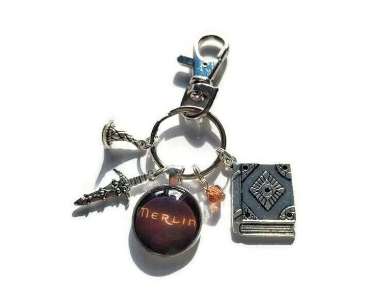 Puede incluir: Un llavero plateado con un dije que dice "Merlin" en una fuente roja y naranja. El llavero tambi&eacute;n tiene un dije de libro plateado, un dije de espada y un peque&ntilde;o dije plateado con un dise&ntilde;o en espiral.