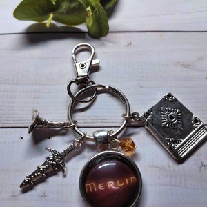 Puede incluir: Un llavero plateado con un dije que dice "MERLIN" en letras doradas sobre un fondo morado oscuro. El llavero tambi&eacute;n tiene un dije de libro plateado, un dije de espada plateado y un dije de trompeta plateado.