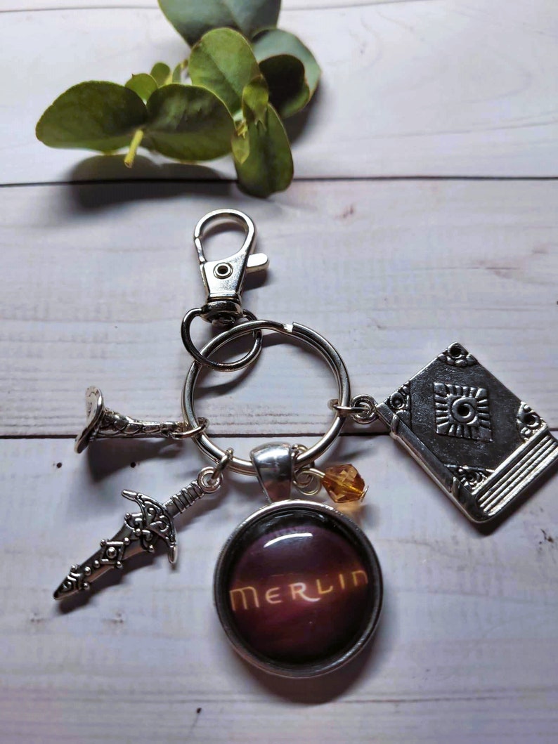 Puede incluir: Un llavero plateado con un dije que dice "MERLIN" en letras doradas sobre un fondo morado oscuro. El llavero tambi&eacute;n tiene un dije de libro plateado, un dije de espada plateado y un dije de trompeta plateado.
