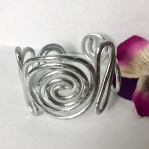 K&ouml;nnte beinhalten: Ein silberfarbenes Manschettenarmband aus Metall mit einer ausgepr&auml;gten geh&auml;mmerten Textur und einem durchgehenden Spiraldesign. Das offene Band besteht aus einem einzelnen dicken Draht, der sich zu einem kreisf&ouml;rmigen Muster windet.