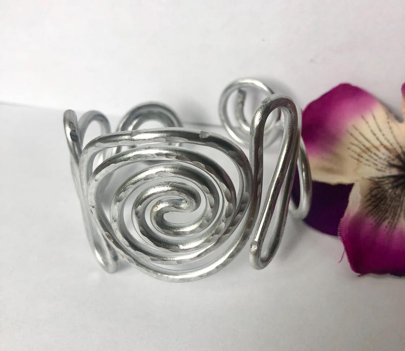 K&ouml;nnte beinhalten: Ein silberfarbenes Manschettenarmband aus Metall mit einer ausgepr&auml;gten geh&auml;mmerten Textur und einem durchgehenden Spiraldesign. Das offene Band besteht aus einem einzelnen dicken Draht, der sich zu einem kreisf&ouml;rmigen Muster windet.