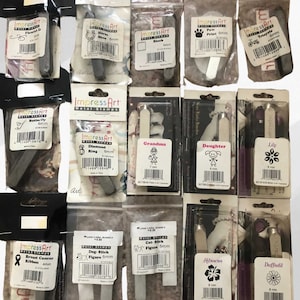 May include: Assortment of metal stamps in individual plastic packaging. The stamps are for crafting and feature various designs, including a butterfly, a diamond ring, a horse shoe, a paw print, a tricycle, a grandma, a daughter, a lily, a breast cancer ribbon, a dog stick, a cat stick, a hibiscus flower, and a daffodil. The stamps are all 6mm in size.