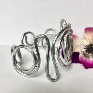 K&ouml;nnte beinhalten: Ein silberfarbenes Metall-Armband in Manschettenform, bestehend aus einer Reihe von miteinander verbundenen, geh&auml;mmerten Drahtspiralen und geschwungenen Schlaufen. Das Armband hat eine polierte, reflektierende Oberfl&auml;che und ein offenes Design.