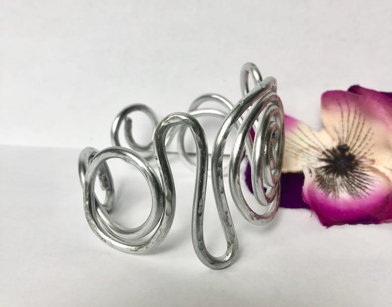 K&ouml;nnte beinhalten: Ein silberfarbenes Metall-Armband in Manschettenform, bestehend aus einer Reihe von miteinander verbundenen, geh&auml;mmerten Drahtspiralen und geschwungenen Schlaufen. Das Armband hat eine polierte, reflektierende Oberfl&auml;che und ein offenes Design.