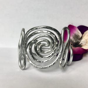 K&ouml;nnte beinhalten: Ein silberfarbenes Manschettenarmband aus Metall mit einem markanten, spiralf&ouml;rmigen Design in der Mitte. Die Metalloberfl&auml;che ist durchgehend geh&auml;mmert und texturiert, was f&uuml;r ein reflektierendes Aussehen sorgt. Das Armband ist offen und mit geschwungenen Drahtdetails versehen.