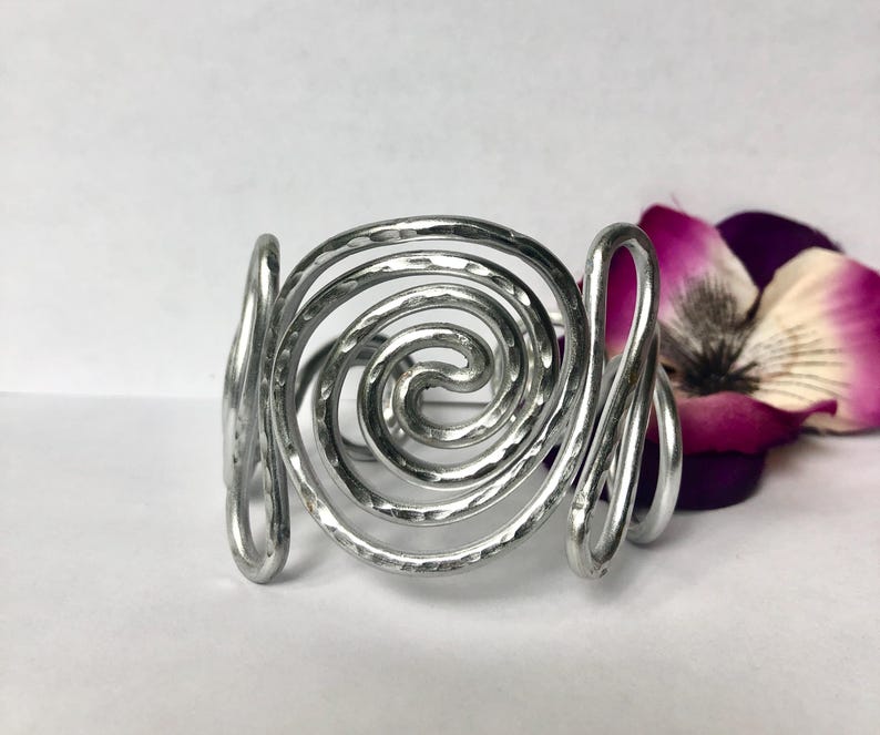 K&ouml;nnte beinhalten: Ein silberfarbenes Manschettenarmband aus Metall mit einem markanten, spiralf&ouml;rmigen Design in der Mitte. Die Metalloberfl&auml;che ist durchgehend geh&auml;mmert und texturiert, was f&uuml;r ein reflektierendes Aussehen sorgt. Das Armband ist offen und mit geschwungenen Drahtdetails versehen.