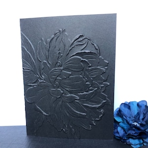 May include: A black card with an embossed peony flower design. The floral image is highly detailed, with visible petals and leaves. The card is standing upright, showcasing the textured surface and the depth of the embossed design. A blue flower is in the background.