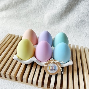 Könnte beinhalten: Sechs pastellfarbene Ostereier in einem weißen Eierhalter aus Keramik, auf einem Holztablett mit Lamellen. Die Eier sind in Rosa, Gelb, Blau und Lila gehalten. Ein kleines Etikett mit einer Zeichnung und dem Schriftzug "Happy Easter" ist angebracht.