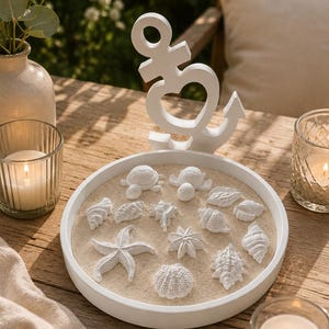 Könnte beinhalten: Ein weißes Ankerornament mit herzförmigem Ausschnitt steht über einem weißen Tablett mit Sand und weißen Muschel- und Seesternfiguren. Zwei brennende Kerzen und eine Blumenvase sind ebenfalls zu sehen.