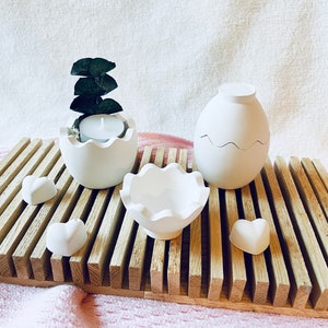Könnte beinhalten: Eine Sammlung weißer Keramik-Dekorationsartikel, darunter eiförmige Behälter, eine Muschelschale und herzförmige Akzente, angeordnet auf einem Holztablett mit Lamellen. Ein eiförmiger Behälter enthält ein brennendes Teelicht und Grünzeug. Die Artikel sind vor einem rosa-weißen Hintergrund platziert.
