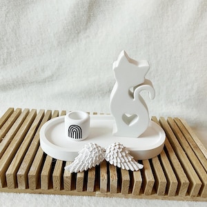 Könnte beinhalten: Eine weiße katzenförmige Figur mit einem Herz-Ausschnitt, ein kleiner Kerzenhalter mit Regenbogenmuster und zwei weiße Engelsflügel werden auf einem weißen ovalen Tablett präsentiert. Das Tablett steht auf einer Holzlattenbasis.