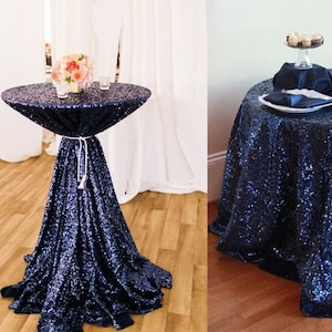 May include: Two navy blue sequin tablecloths, one draped over a round cocktail table and the other on a round dining table. Both tablecloths have a sparkly, iridescent finish.
