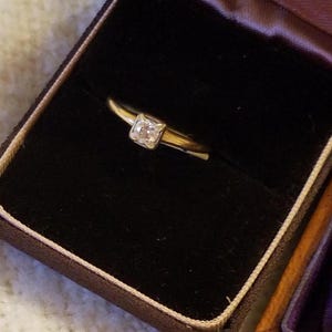 May include: A gold engagement ring with a square-shaped diamond in a black velvet ring box.