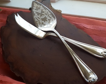 May include: A silver cake server and butter knife set on a wooden serving board. The server has an ornate design and the handles are both silver.