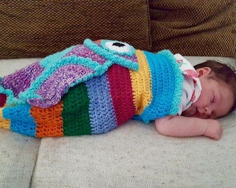 May include: A baby sleeping in a colorful crocheted fish-shaped swaddle blanket. The blanket is striped with blue, green, yellow, orange, and red.