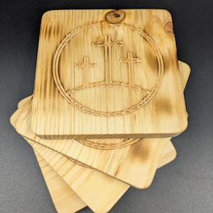 May include: A stack of square wooden coasters with a carved design. Each coaster features a circle with three crosses, likely representing a religious theme. The wood has a light, natural color with some darker, burnt accents.