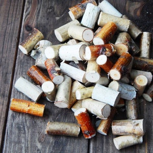 Może przedstawiać: Stos małych, białych i brązowych drewnianych patyczków, niektóre z naturalną fakturą kory, na ciemnobrązowej drewnianej powierzchni.
