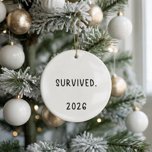 May include: A white ceramic Christmas ornament with the text "SURVIVED. 2026" in black. The ornament hangs from a gold string against a backdrop of a decorated Christmas tree with white and gold ornaments.