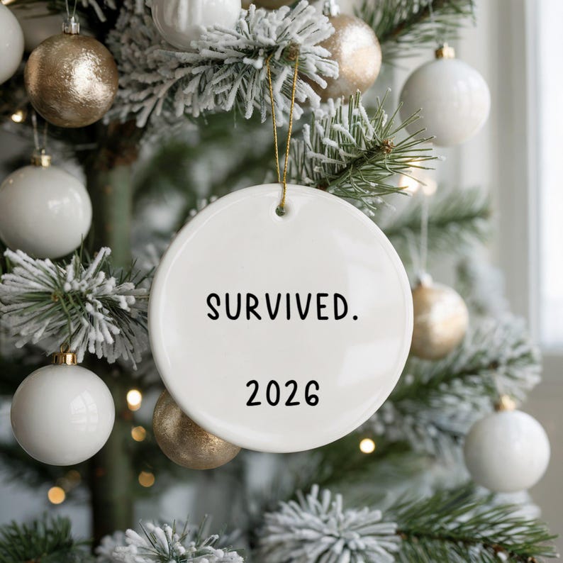 May include: A white ceramic Christmas ornament with the text "SURVIVED. 2026" in black. The ornament hangs from a gold string against a backdrop of a decorated Christmas tree with white and gold ornaments.