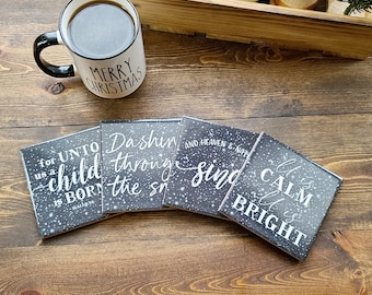 May include: Four black square coasters with white text and a snowy background. The text on the coasters reads: "for unto us a child is born", "Dashing through the snow", "And heaven & earth sing", and "All is calm all is bright".