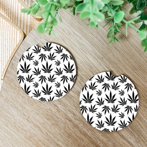 May include: Two round car coasters with a white background and a repeating black cannabis leaf pattern. The coasters have a small notch for easy removal from the car's cup holder. The coasters are on a wooden surface.