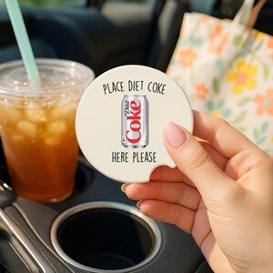 May include: A round, off-white car coaster with the words "PLACE DIET COKE HERE PLEASE" printed in black. The coaster features a graphic of a Diet Coke can. A clear plastic cup with iced tea and a straw is in the background.