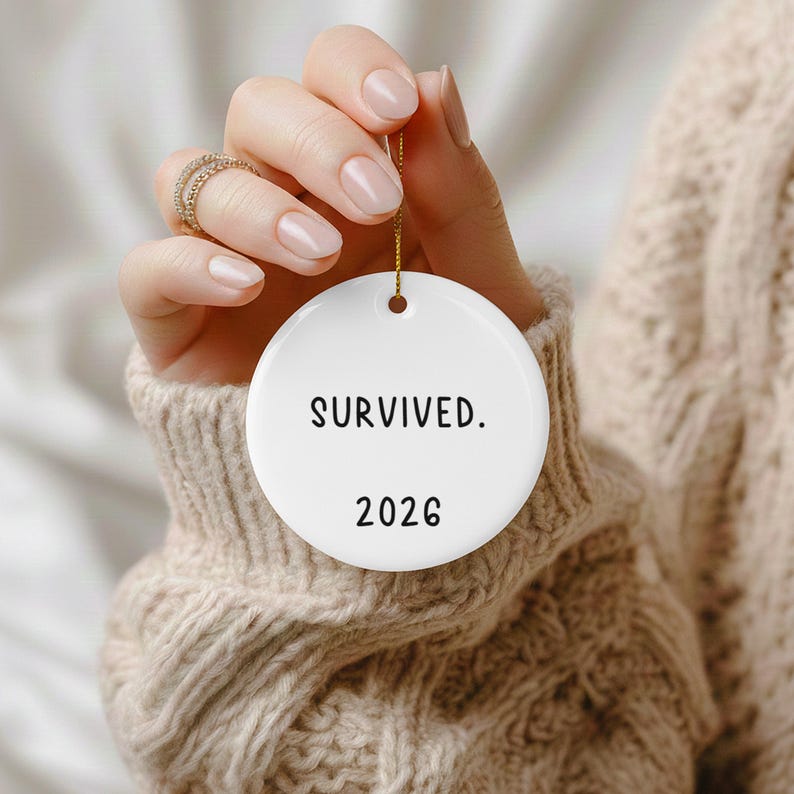 May include: A white, round ornament with the words "SURVIVED." and the year "2026" printed in black. The ornament is suspended by a gold string. The background is a soft, neutral color.