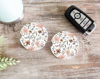 May include: Two round car coasters with a floral pattern. The coasters feature a white background with brown and pink watercolor flowers and leaves.
