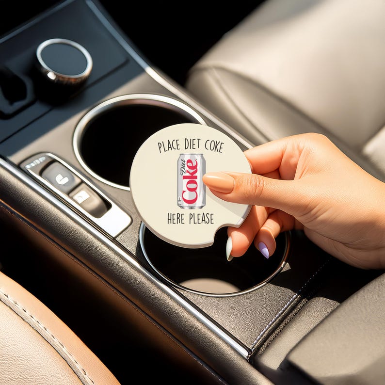 May include: A round, beige car coaster with the text "Place Diet Coke Here Please" and a Diet Coke can graphic. The coaster is being held in a car's cup holder.